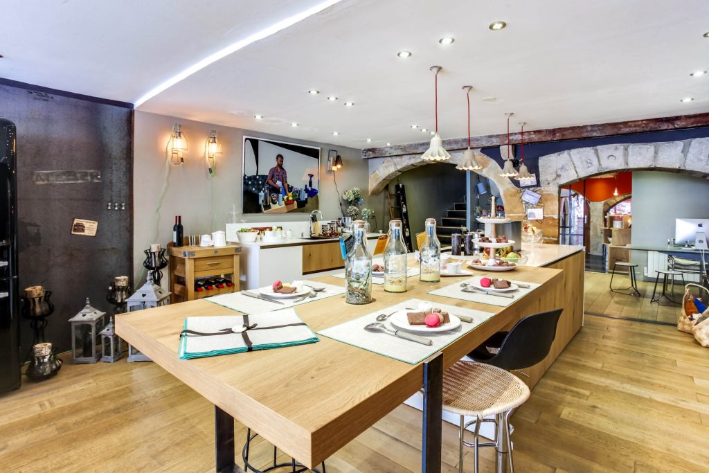 Nouvelle expo cuisines chez Bulles Cuisines, à Lyon. Îlot central bois et blanc laqué avec plaque de cuisson induction intégrée, hotte design escamotable, et table de repas style plan de travail sur pieds métalliques. Zoom sur l’îlot combiné avec la table bois.