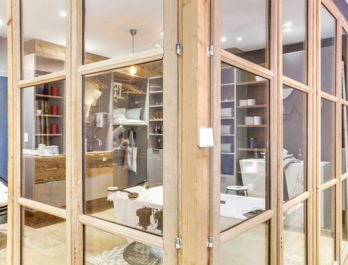 Univers salle de bain chez Bulles Cuisines à Lyon. Vue à travers une cloison vitrée : salle de bain avec baignoire, dressing et 2 meubles vasques XXL au design contemporain.