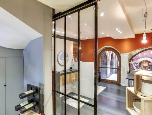 Univers salle de bain chez Bulles Cuisines à Lyon. Salle d'eau avec douche italienne, paroi vitrée, mur effet bois, sèche serviette chromé effet miroir. Meuble bois et blanc laqué, meuble vasque contemporain en pierre anthracite. Verrière type atelier.