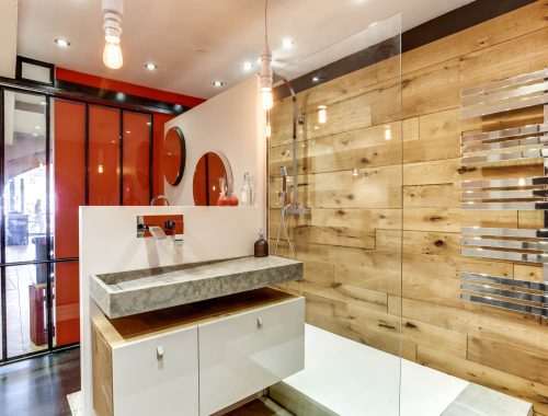 Univers salle de bain chez Bulles Cuisines à Lyon. Salle d'eau avec douche italienne, paroi vitrée, mur effet bois, sèche serviette chromé effet miroir. Meuble bois et blanc laqué, double vasque pierre. Verrière type atelier et pan de mur orange vitaminé.